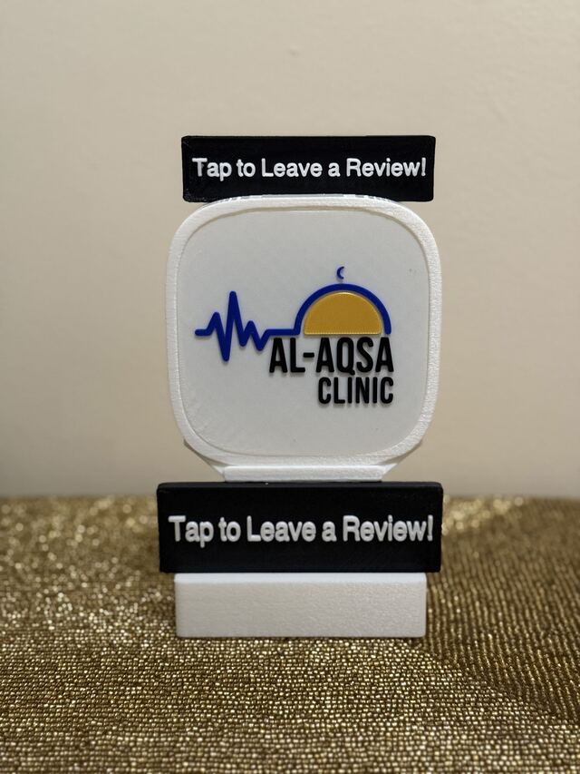 A white TapCraft Stand Pro with the Al-Aqsa Community Clinic logo and 'Tap to Leave a Review!' signs top and bottom.