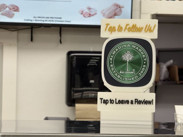 A TapCraft Stand Pro at Almadina Market with two signs — 'Tap to Follow Us!' on top and 'Tap to Leave a Review!' below — in front of the menu boards.