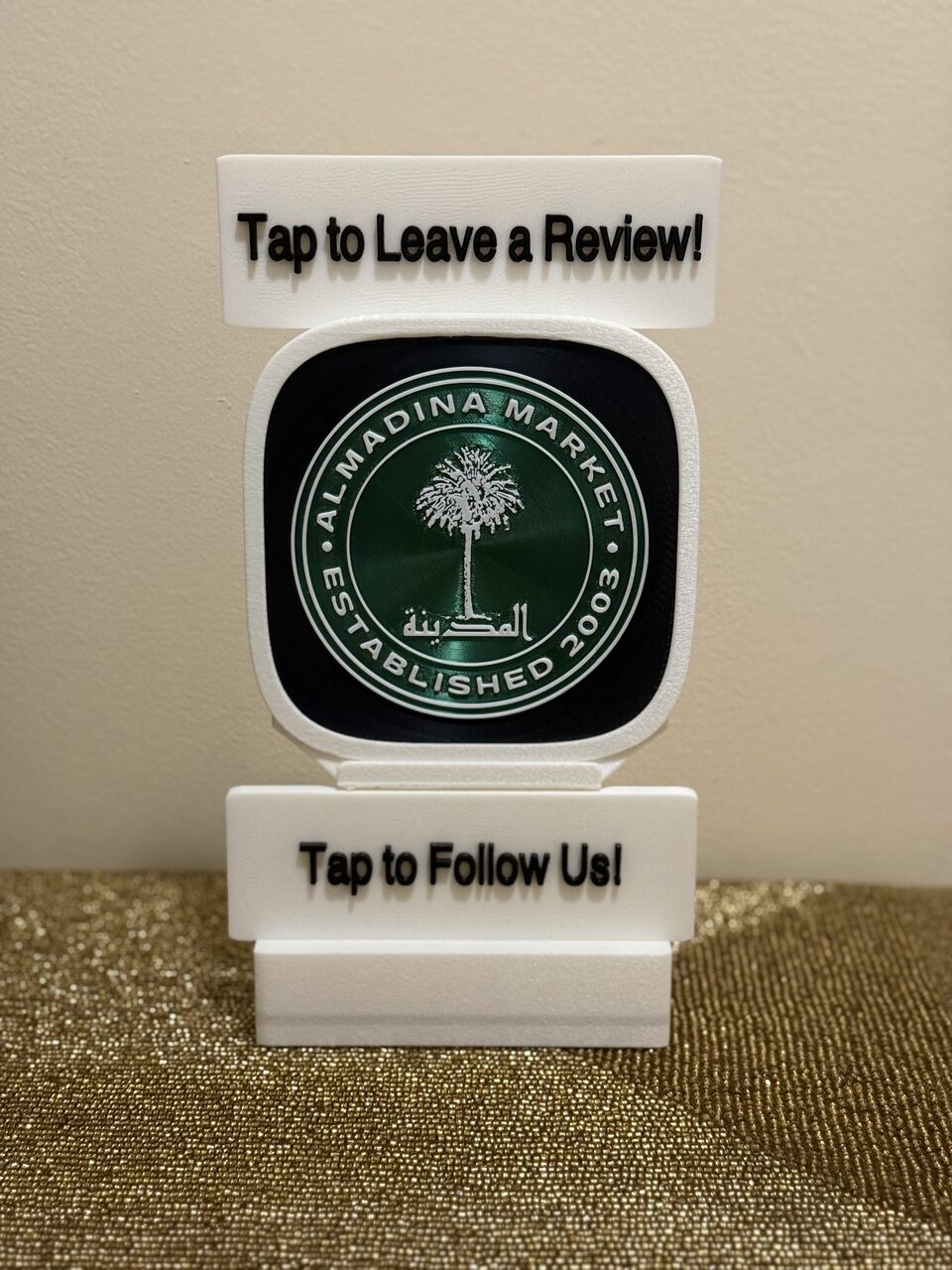 A TapCraft Stand Pro at Almadina Market. Two signs frame the NFC display — 'Tap to Leave a Review!' on top and 'Tap to Follow Us!' on the bottom.